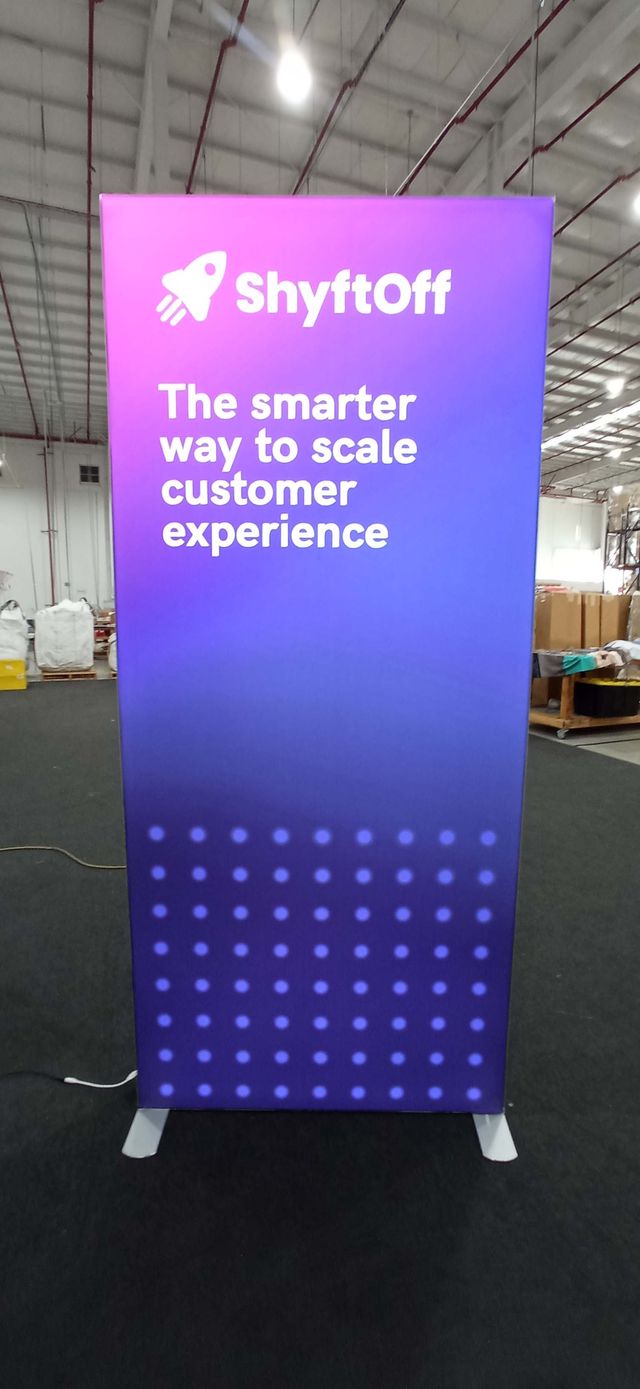 A tall, rectangular illuminated lightbox sign set up on a floor. The sign has a purple gradient background with white text.