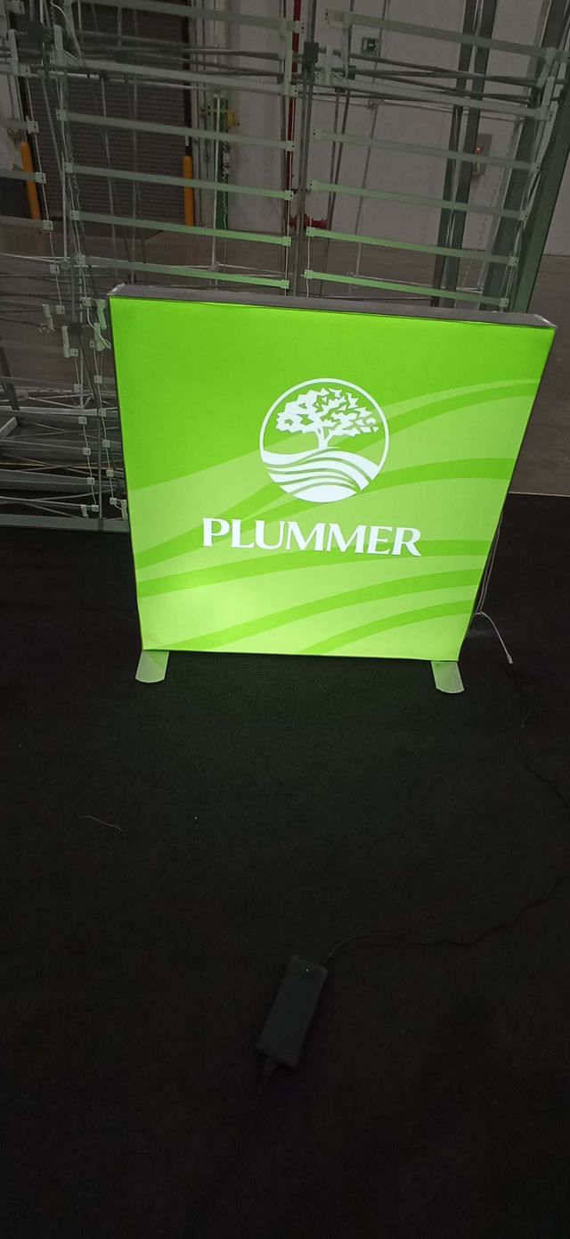 A small, square illuminated lightbox sign set up on a floor. The sign is green with a logo and text in white.