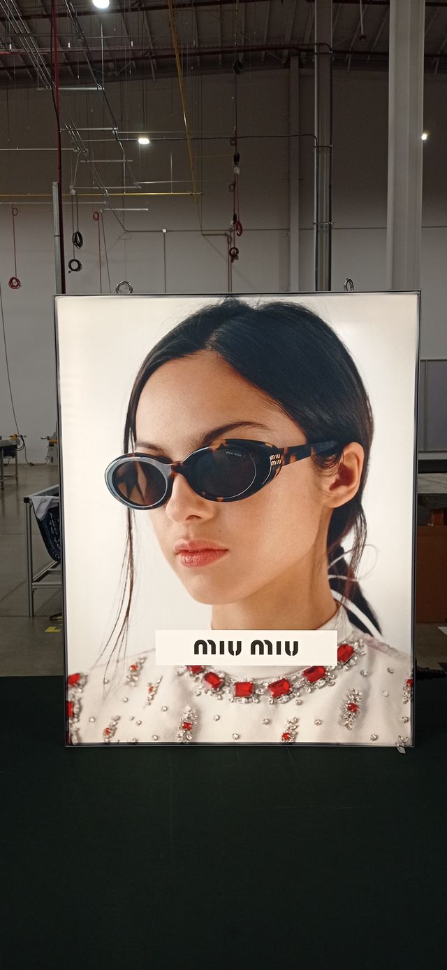 An illuminated display showing a woman wearing sunglasses and a shirt with red jewels on it. The brand Miu Miu is at the bottom.